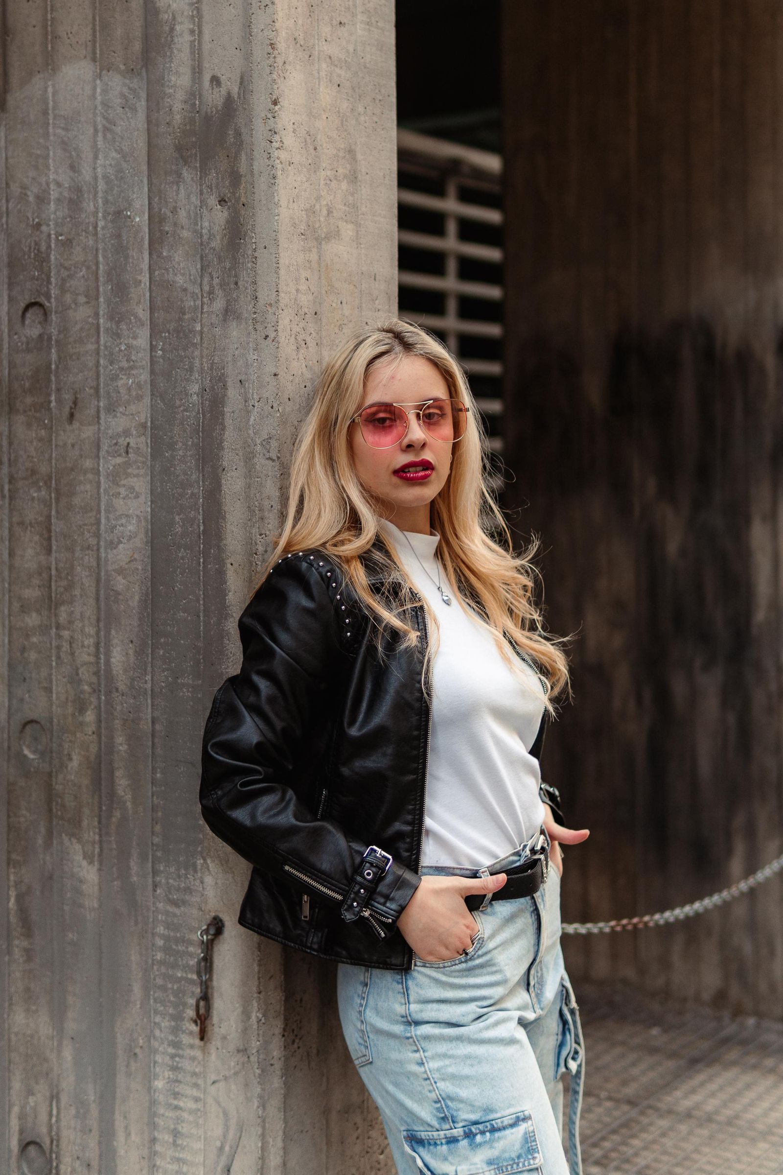 Martina con chaqueta negra y gafas rosadas junto a una pared de hormigón.