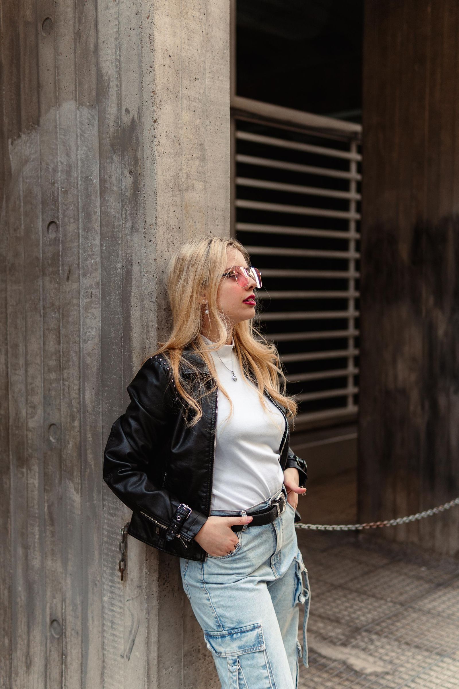 Martina posa de cuerpo entero con chaqueta negra, jean y gafas rosadas junto a una pared de hormigón.